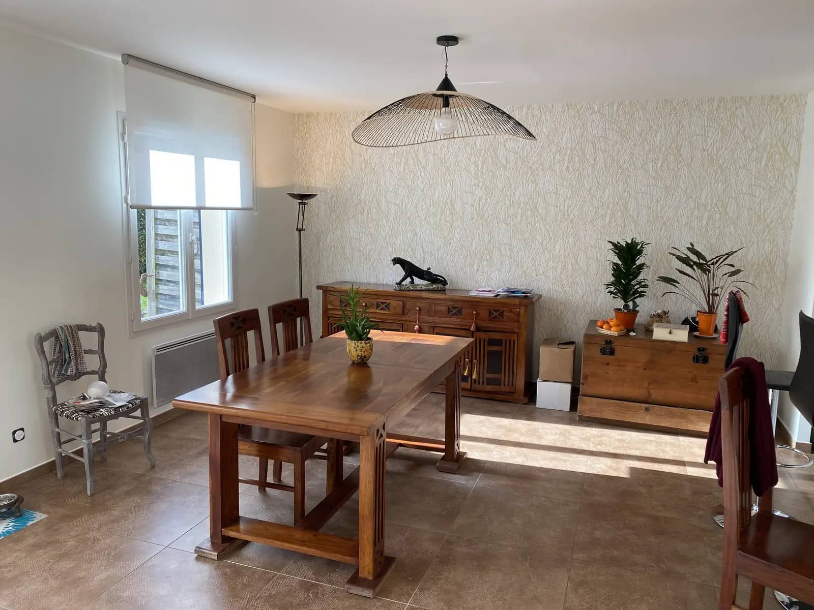 Photo d'une salle à manger. Le mur principal est recouvert d'un papier peint beige clair à motif linéaire évoquant des herbes hautes, créant une ambiance naturelle et sereine.