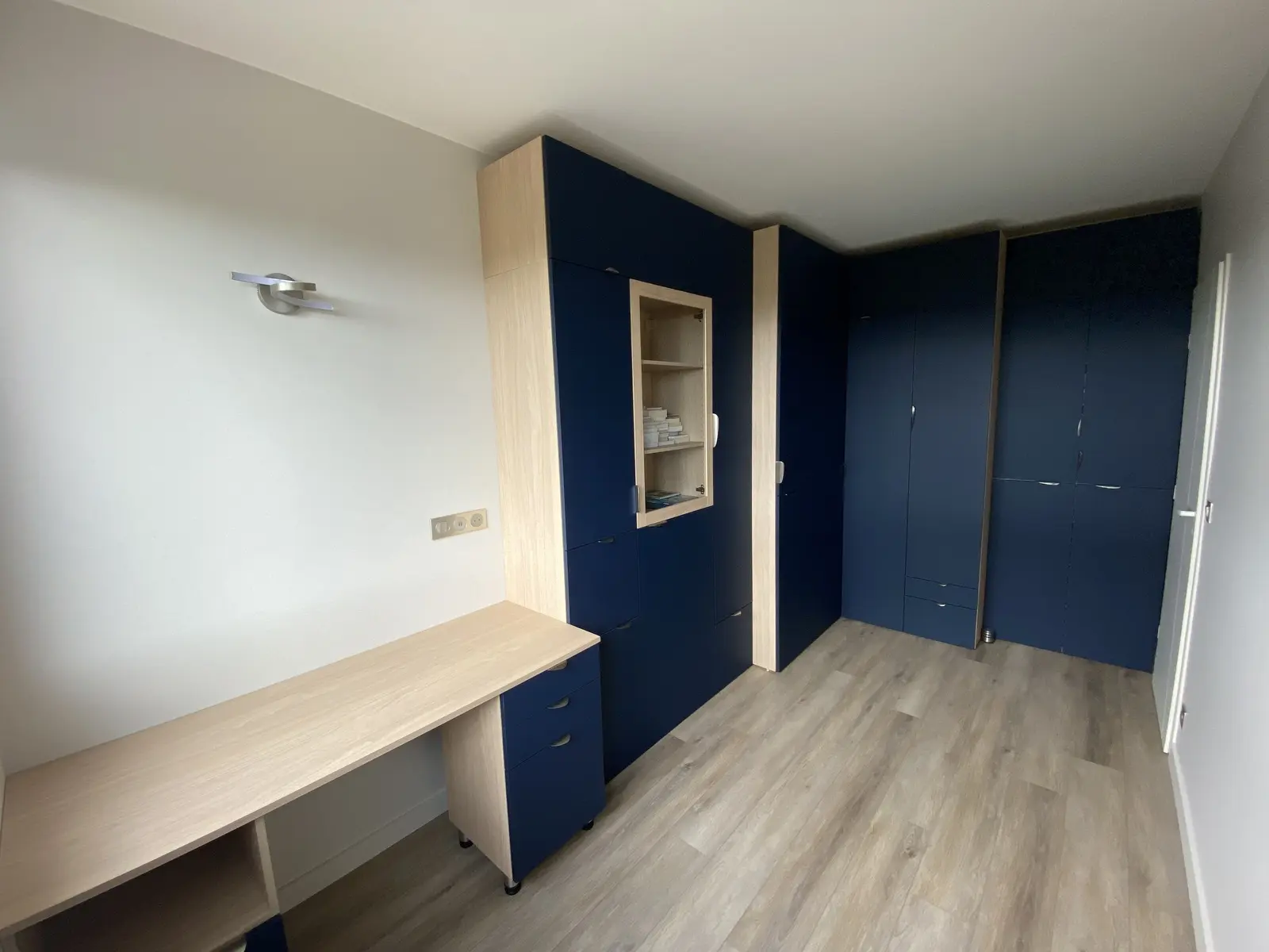 Photo d'un aménagement intérieur sur mesure. Des meubles aux couleurs bleu foncé et bois clair sont disposés dans une pièce. On observe un bureau, des armoires murales et une disposition optimisant l'espace.