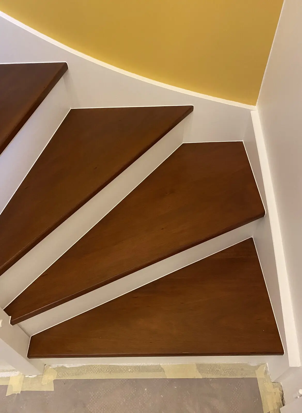 Escalier en bois récemment rénové. Les marches sont en bois brun foncé, avec des contremarches blanches. Du ruban de masquage est visible au sol, suggérant que des travaux de peinture ou de finition sont en cours ou viennent d'être terminés. L'accent est mis sur le bois riche et la netteté des lignes, mettant en valeur la qualité de la rénovation.