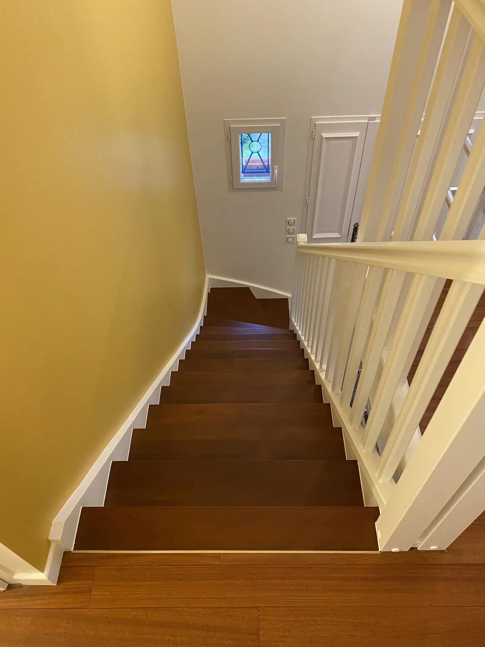Escalier rénové, vu du haut. Les marches sont en bois brun foncé, et la rampe est blanche. La rénovation met en valeur le contraste entre le bois foncé et le blanc de la rampe, ainsi que la couleur vive du mur. L'ensemble donne une impression de modernité et de propreté.