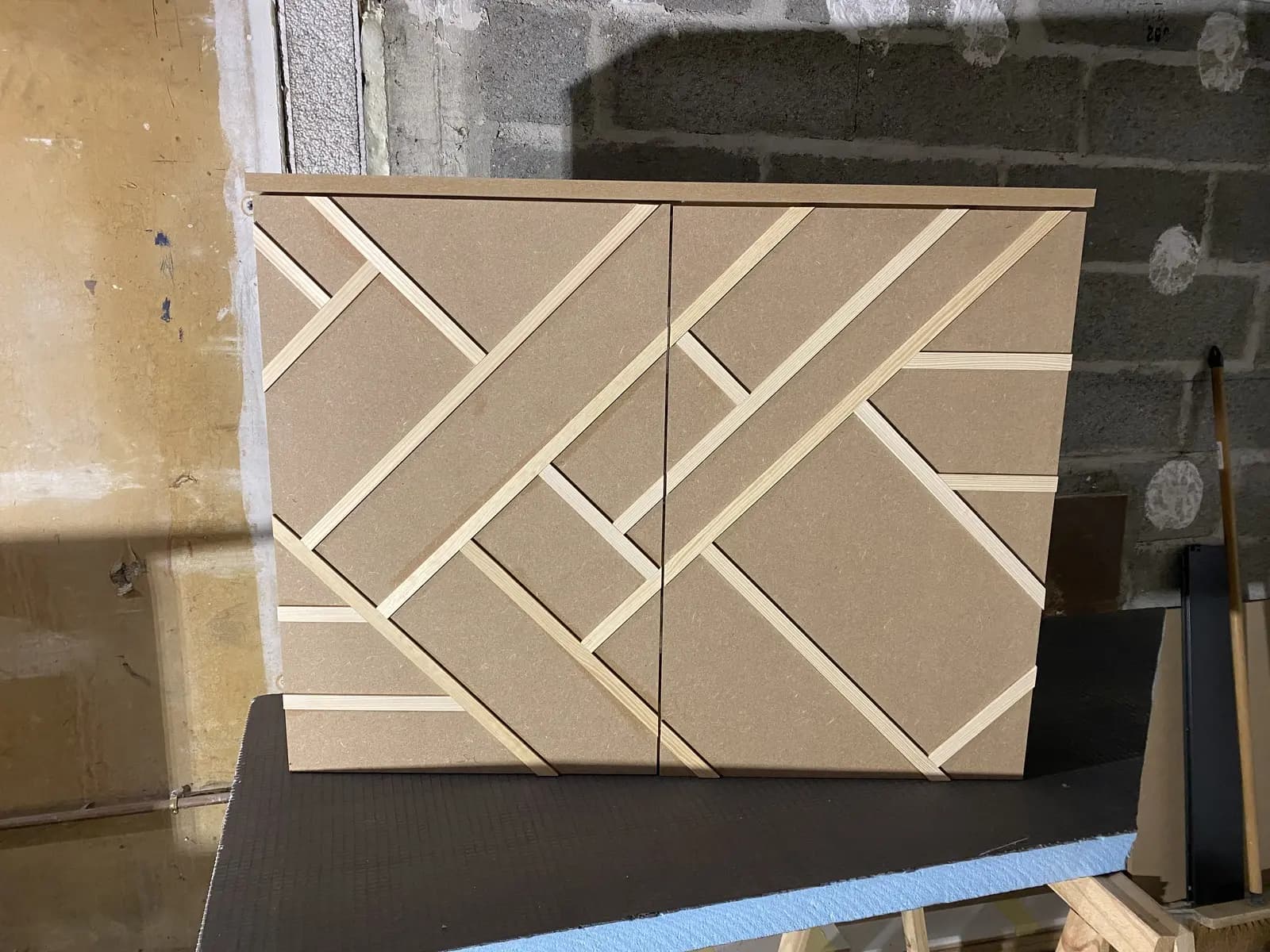 Placard sur mesure en phase de construction. Deux panneaux de particules beige clair sont assemblés, décorés d'un motif géométrique composé de lignes de bois clair disposées en diagonale, créant un motif en losanges. Le meuble est posé sur un sol avec des matériaux de construction.