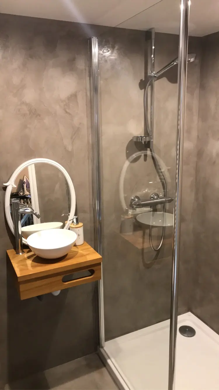 Salle de bain dont les murs sont entièrement recouverts d'un enduit décoratif gris, dont les nuances subtiles et les textures légèrement irrégulières apportent une profondeur visuelle et un aspect artisanal très élégant. Une cabine de douche en verre, un lavabo en céramique blanche sur un meuble en bois clair, et un miroir rond complètent la pièce.
