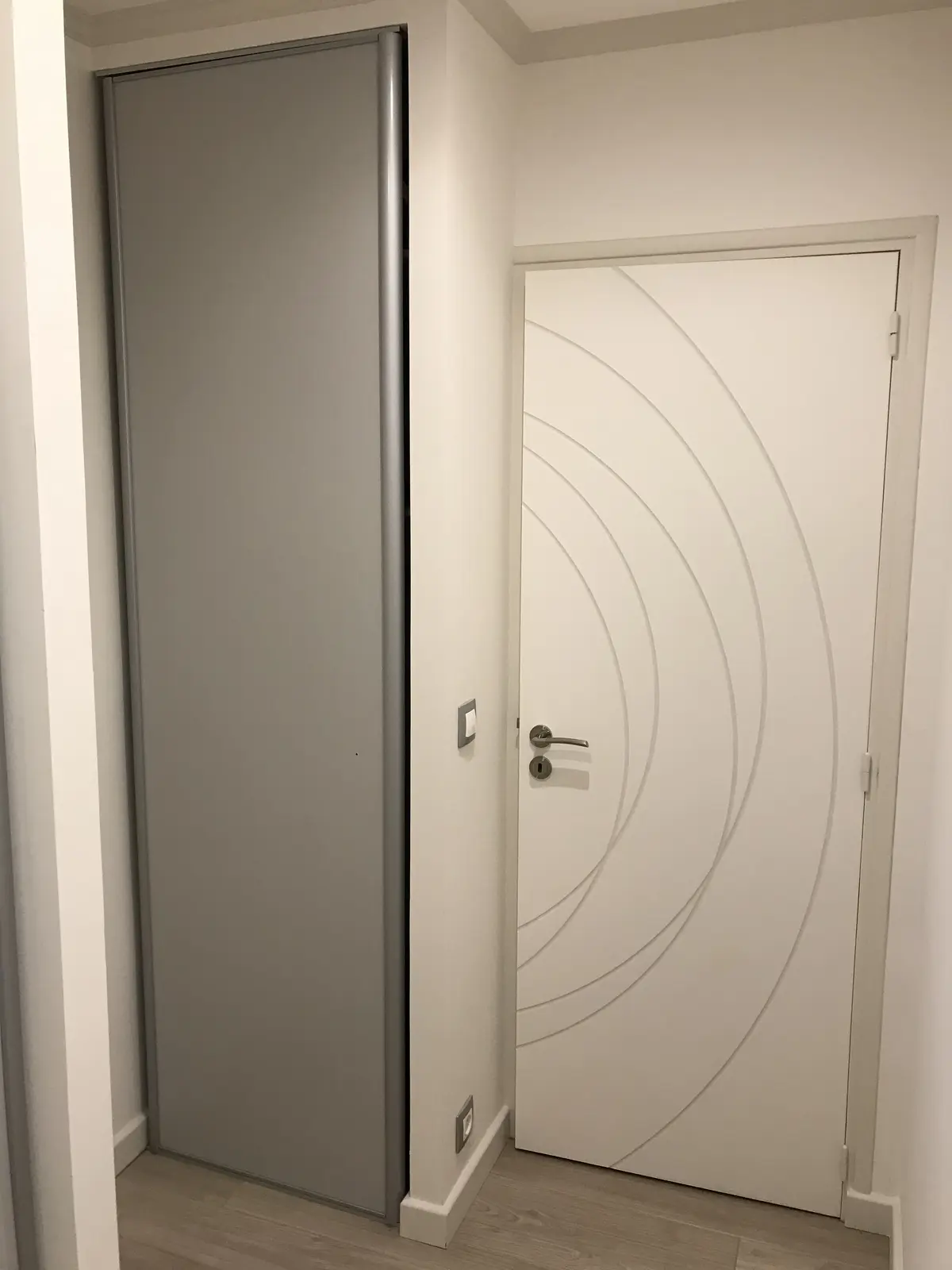 Couloir rénové avec porte blanche moderne : lignes courbes en relief aux rainures soulignées d’une teinte foncée. Plinthes de plafond et encadrement assortis, créant une harmonie élégante et contemporaine.
